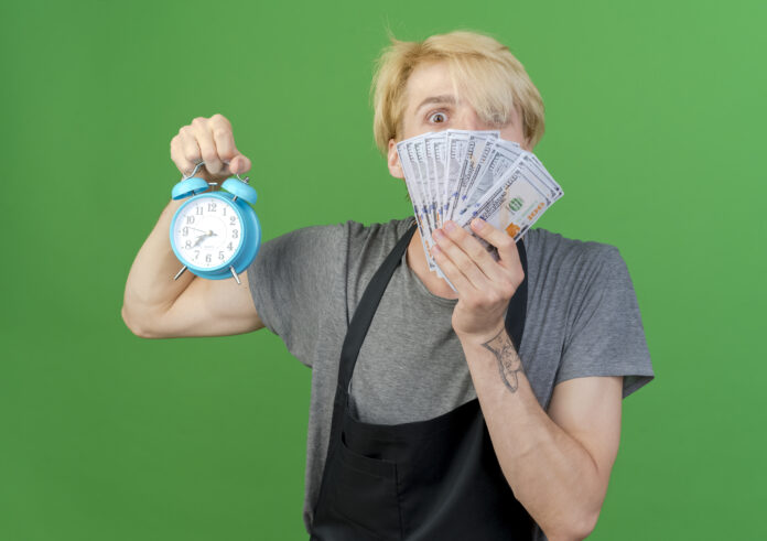professional barber man in apron alarm clock and cash looking at camera surprised standing over green background
